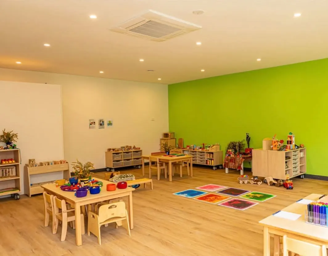 Bright learning room at YoMySunshine with child-sized timber tables, open shelves of toys and a green feature wall