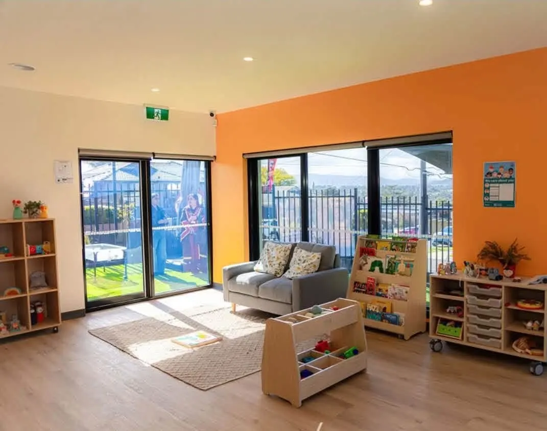 Sunny play room with a grey couch, rug, picture-book display and low toy shelves beside large windows