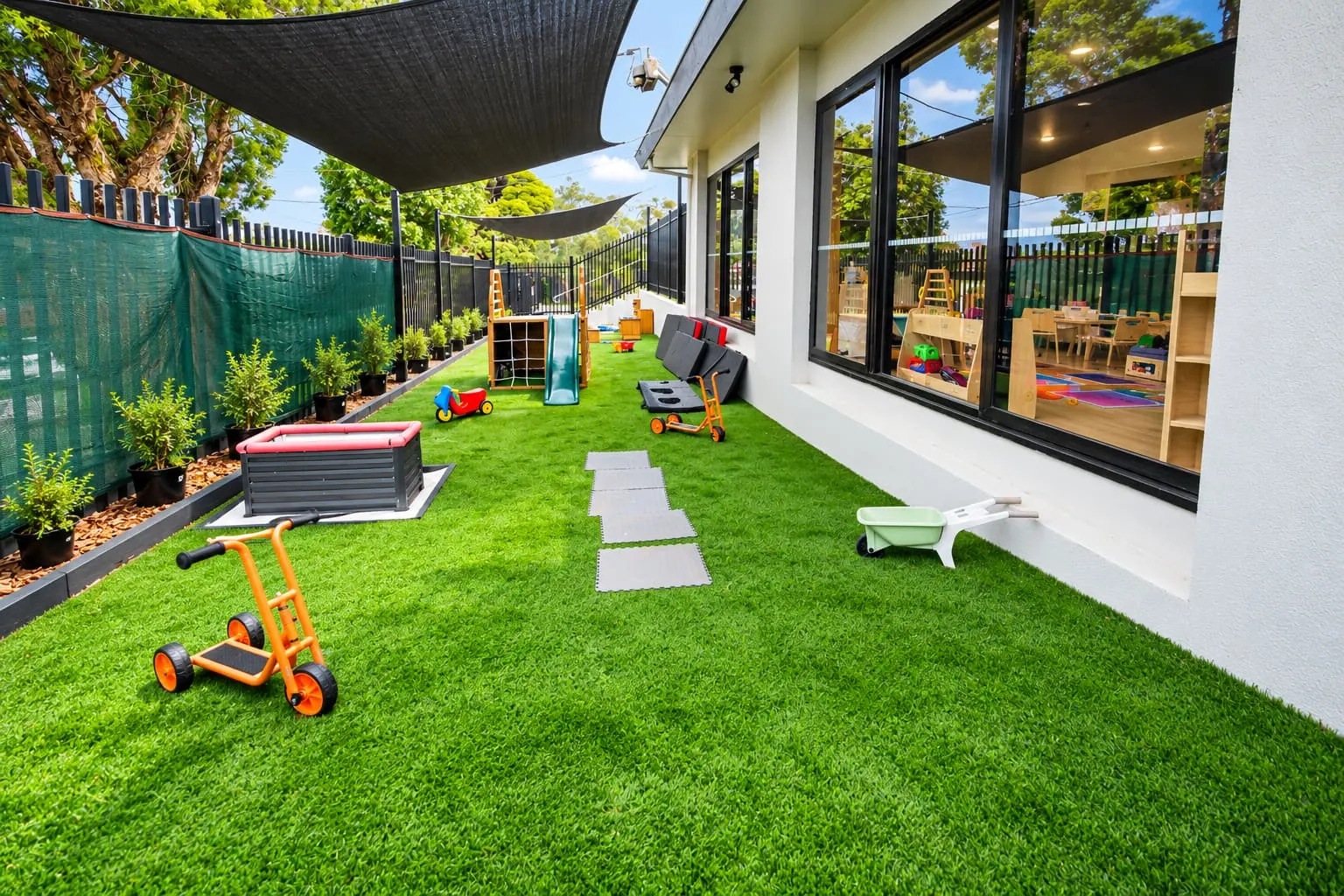Outdoor play space beside the centre, with ride-on toys on artificial grass under shade sails