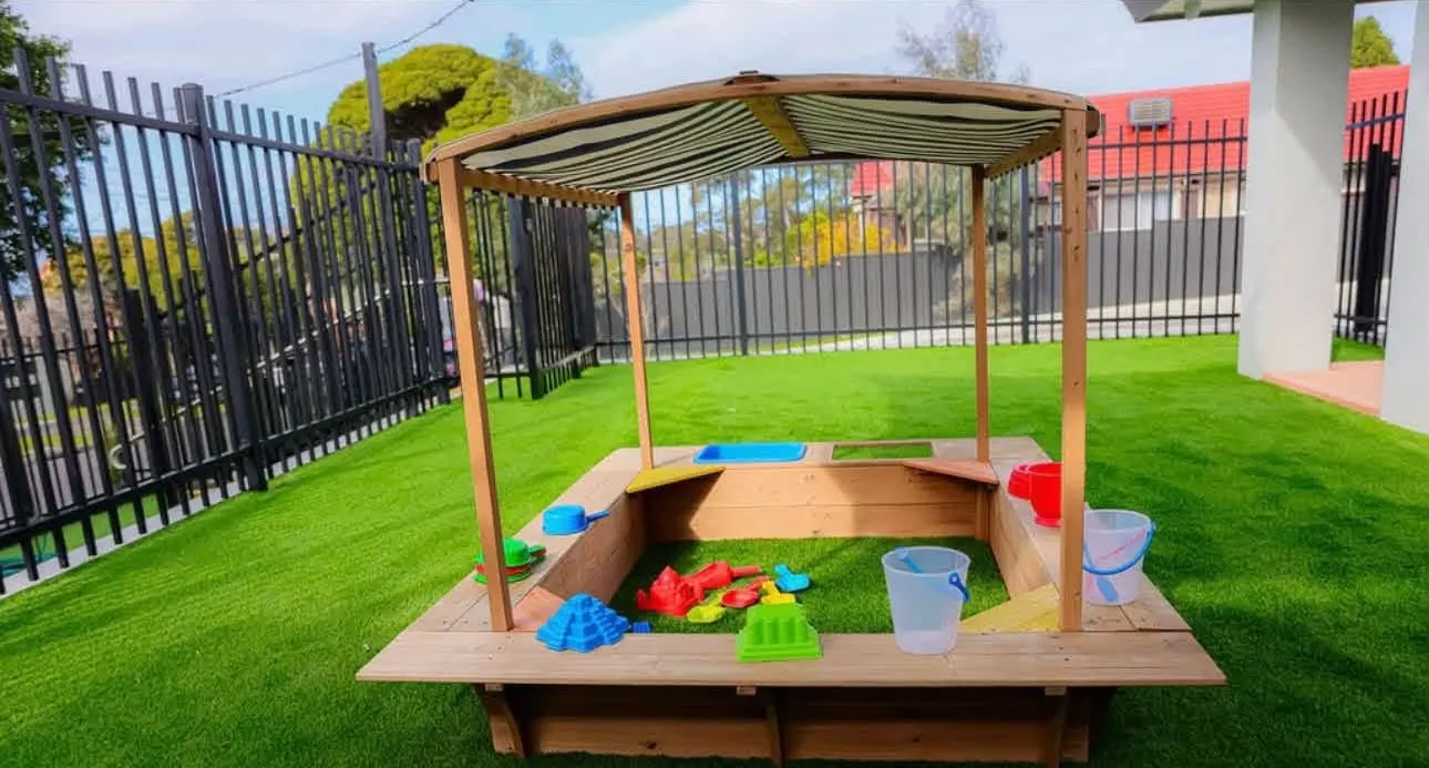 Covered timber sandpit with buckets, spades and moulds on synthetic grass inside a fenced play area